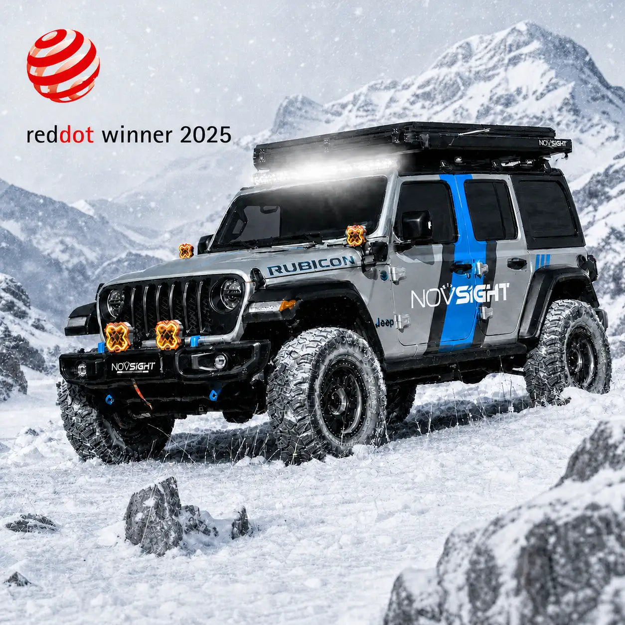 A silver and blue off-road Jeep Wrangler parked in a snowy mountain landscape, with a roof rack, front bumper kit, and off-road tires. The image includes a 'reddot winner 2025' badge in the top-left corner.