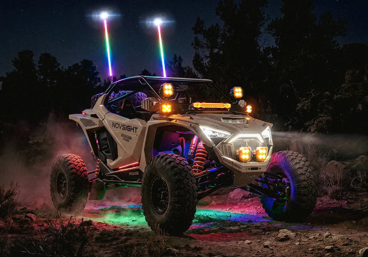 A white Canopy Novysight UTV off-road vehicle at night with colorful LED underglow and roof-mounted light bars, driving on a dirt trail with trees in the background.