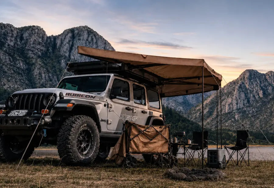 A white Rubicon off-road vehicle parked outdoors in a mountain landscape at sunset, with a tan rooftop awning extended over the side and camping chairs and a storage bag set up beneath it.