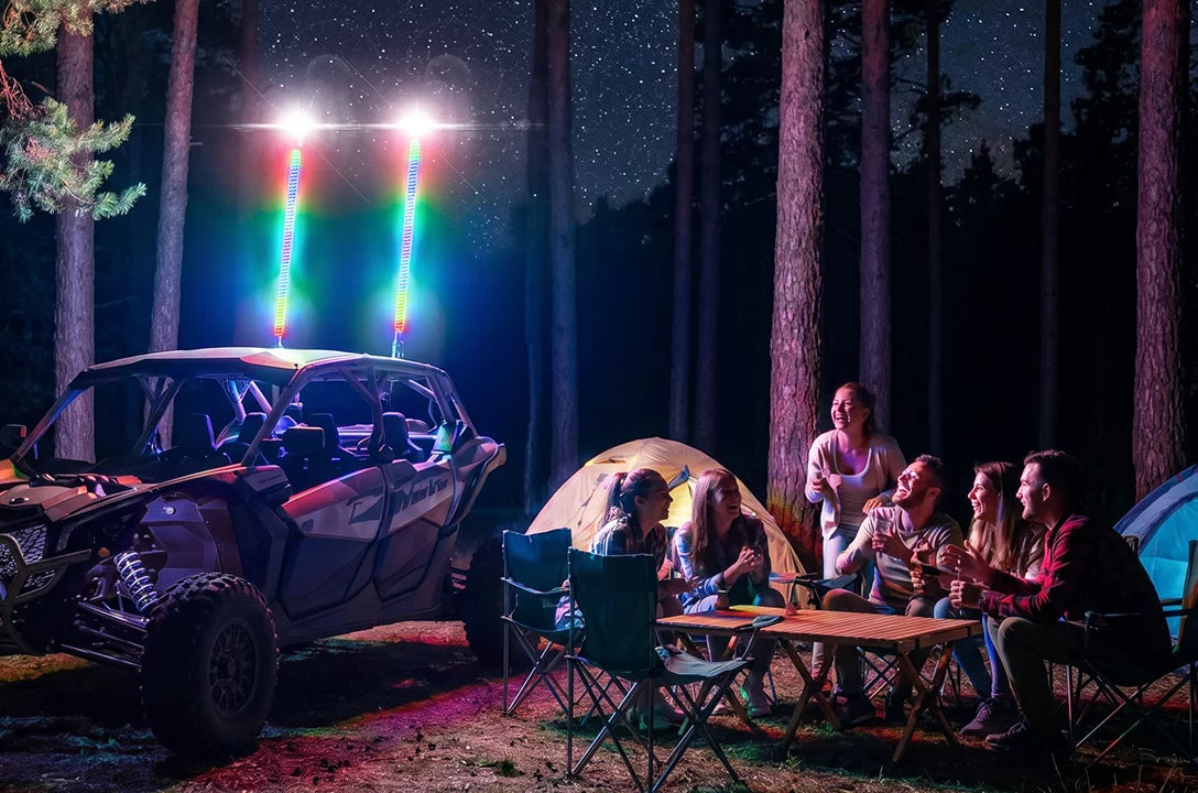 A group of friends camping at night in a pine forest, sitting around a small table with a tent and a side-by-side vehicle with bright multicolor light bars mounted on the roof. The scene is illuminated by the colorful lights against a dark starry sky.