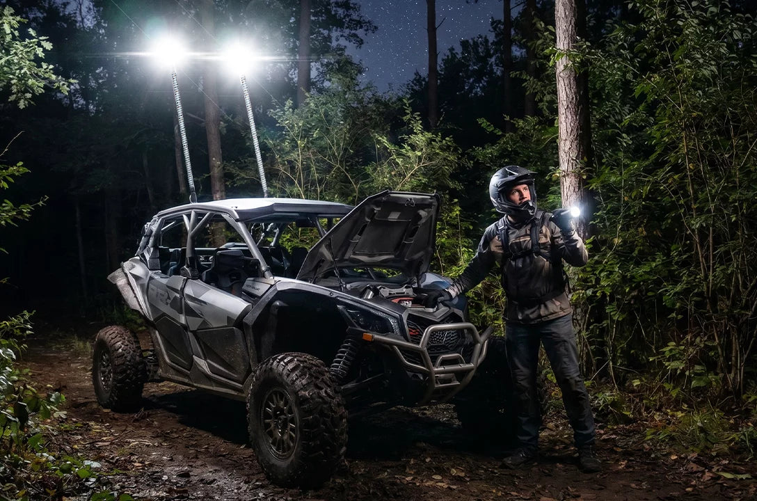 A rugged off-road vehicle with its hood open stands on a forest trail at night, illuminated by bright vehicle lights and a person in full riding gear holding a flashlight beside it.