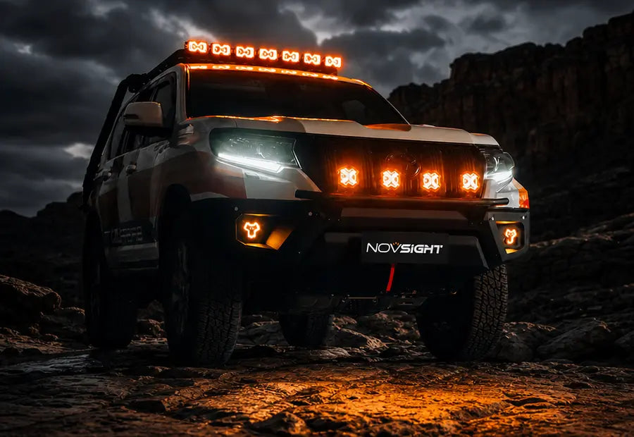 A rugged off-road SUV with illuminated amber LED light bars and front fog lights, parked on rocky terrain at night under a dark cloudy sky. The front bumper shows the brand name NOVSIGHT.
